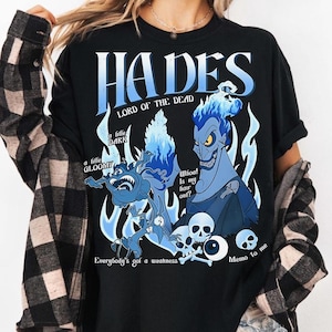 Può includere: T-shirt nera con grafica blu di Ade di Hercules della Disney. Il design include il testo "HADES LORD OF THE DEAD" e citazioni come "Whoa! Is my hair out?" e "Everybody's got a weakness."