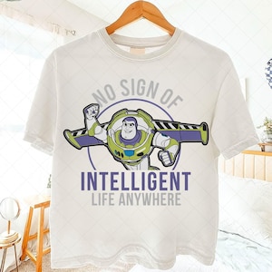 May include: White t-shirt featuring a graphic of Buzz Lightyear with the text "NO SIGN OF INTELLIGENT LIFE ANYWHERE" in purple and gray. The design includes Buzz in his spacesuit, with green, purple, and white accents.