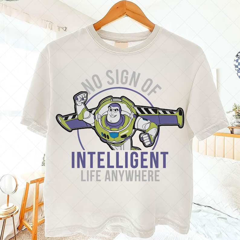 May include: White t-shirt featuring a graphic of Buzz Lightyear with the text "NO SIGN OF INTELLIGENT LIFE ANYWHERE" in purple and gray. The design includes Buzz in his spacesuit, with green, purple, and white accents.