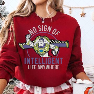 May include: A red sweatshirt featuring a graphic of Buzz Lightyear with the text "NO SIGN OF INTELLIGENT LIFE ANYWHERE". The Buzz Lightyear graphic is in green, white, blue, and purple.