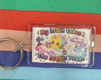 May include: A rectangular clear acrylic keychain featuring a colorful illustration of a smiling, rainbow-colored character reading a book. The text reads, My brain takes the scenic route, surrounded by small hearts, stars, and a winding path.