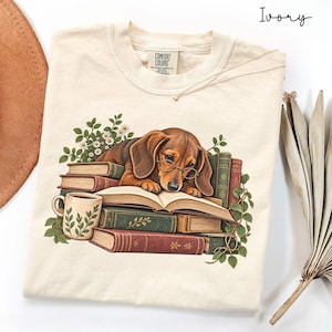 Puede incluir: Una camiseta color marfil con el gráfico de un perro salchicha marrón con gafas redondas, descansando sobre una pila de libros antiguos. La ilustración incluye una taza de cerámica con patrón de hojas, margaritas blancas y enredaderas.