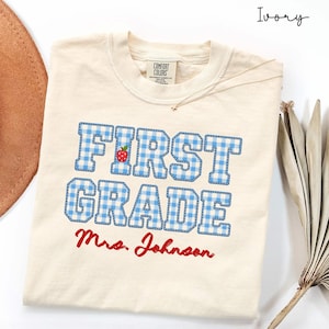 May include: An ivory t-shirt featuring the text FIRST GRADE in blue and white gingham block letters with a small red apple graphic inside the letter R. Below the text, the name Mrs. Johnson is printed in red cursive script.