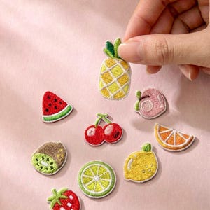 Può includere: Toppe ricamate con frutta, tra cui ananas, fetta di anguria, ciliegie, kiwi, limone, fetta d'arancia, fragola, pesca e lime. Le toppe sono colorate con cuciture bianche su sfondo rosa.