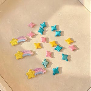 May include: A collection of embroidered fabric patches featuring three yellow shooting stars with pink and blue trails, alongside several small four-pointed star shapes in assorted colors including bright yellow, light pink, and vibrant turquoise blue.