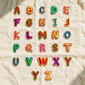 May include: A collection of colorful, embroidered alphabet patches. Each letter is a different color, with a white border. The letters are arranged in rows and columns on a white fabric background.