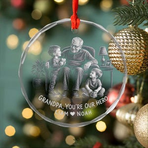 May include: A clear, circular glass ornament featuring an etched illustration of a grandfather sitting in a chair with two young children. The text reads, "GRANDPA, YOU'RE OUR HERO" followed by the names "LIAM" and "NOAH" with a heart symbol.