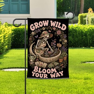 Könnte beinhalten: Eine schwarze Gartenflagge mit einem Skelett in einem geblümten Hut und einer Schürze, das in einem halbmondförmigen Pflanzgefäß gärtnert. Der Text "GROW WILD" und "BLOOM YOUR WAY" steht in rosa Buchstaben zwischen Blumen und Gartenwerkzeugen.