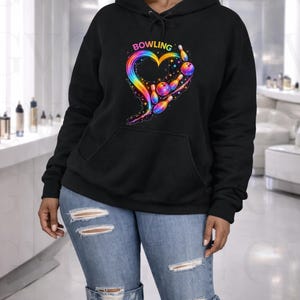 Puede incluir: Sudadera con capucha negra con bolsillo delantero y capucha con cordón. La sudadera presenta un diseño en forma de corazón colorido con bolos y bolas de bolos, y la palabra "BOWLING" encima del corazón. El diseño incorpora colores del arcoíris.