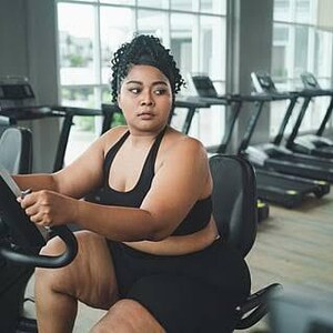 May include: A person with dark curly hair tied back is wearing a black athletic sports bra and matching black shorts while sitting on a stationary exercise bike in a gym setting with multiple treadmills visible in the background.