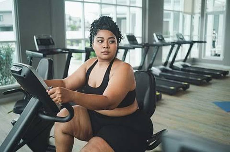 May include: A person with dark curly hair tied back is wearing a black athletic sports bra and matching black shorts while sitting on a stationary exercise bike in a gym setting with multiple treadmills visible in the background.