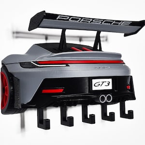 May include: A silver Porsche GT3 wall-mounted key holder featuring a large black rear spoiler with the word PORSCHE printed on it. The rear of the car model includes red taillights, a black bumper, dual exhaust pipes, and five black hooks.