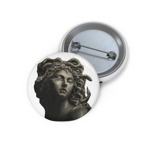 May include: A round pin-back button featuring a black and white illustration of a classical figure with snakes for hair. The design shows a face with closed eyes and horizontal lines across the eyelids. The back of the button is silver metal.