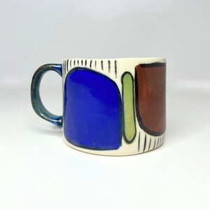 May include: A ceramic mug with a white base, blue handle, and abstract shapes. The mug features blue, brown, and green designs with black vertical lines. It's a standard size, suitable for tea or coffee, with measurements in cm.