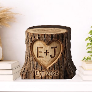 Puede incluir: Un tocón de árbol de madera con un tallado en forma de corazón. El corazón contiene las iniciales "E+J" y el año "EST 2026". El tocón tiene una textura de corteza natural, probablemente un objeto decorativo o un regalo personalizado.