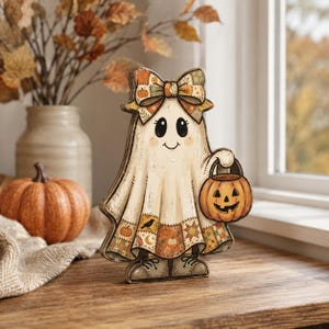 May include: A decorative wooden cutout of a ghost featuring a cream-coloured body, a large bow on its head, and a hemline decorated with a patchwork pattern of pumpkins, stars, and a black bird. The ghost holds a small orange jack-o'-lantern bucket.