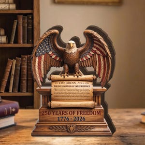 May include: A wooden sculpture of an eagle with outstretched wings, featuring the American flag design on the wings. The sculpture includes a scroll with text from the Declaration of Independence and the text "250 YEARS OF FREEDOM 1776-2026".
