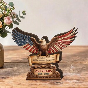 May include: A wooden sculpture of an American bald eagle with outstretched wings, featuring the colours of the American flag. The sculpture includes the text "IN CONGRESS, JULY 4, 1776, The Declaration of Independence" and "250 YEARS 1776-2026".