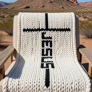 May include: A chunky knit blanket in cream with a large black cross design and the word "JESUS" vertically centered. The blanket is draped over a wooden chair, set against a desert landscape with a mountain backdrop.