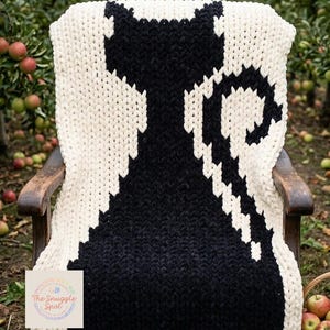 May include: A chunky knit blanket draped over a wooden chair, featuring a large black cat silhouette against an off-white background. The blanket is made of thick yarn, creating a cozy texture. The background shows an orchard with apples.