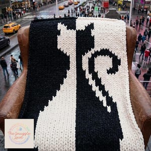 May include: A black and white knitted blanket featuring a cat silhouette design. The blanket is draped over a brown leather chair. The text on the image says "The Snuggle Spot" and "This photo is AI generated and is for illustration purposes only to visualize finished product."