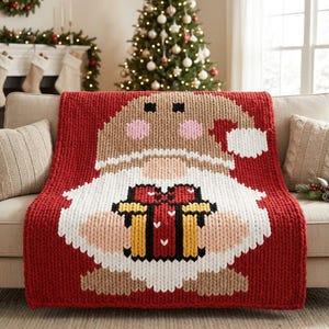 May include: A chunky knit throw blanket featuring a large gnome character centered on a red background. The gnome wears a tan hat with a white pom-pom and holds a gift box with yellow, black, and red details. The design uses thick yarn stitches.