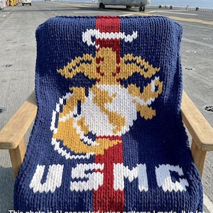 May include: A navy blue knitted blanket featuring the USMC emblem in white, gold, and red. The blanket is draped over a wooden chair on a ship deck, with a military aircraft in the background. The text "USMC" is displayed prominently.