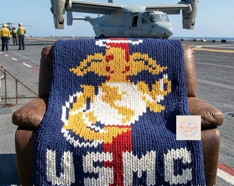 May include: A chunky knit blanket featuring the United States Marine Corps emblem in yellow, white, and red on a dark blue background, with the letters USMC in white at the bottom. The blanket is draped over a brown leather armchair.