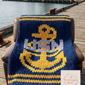 May include: A navy blue knitted blanket featuring a large anchor design in yellow, white, and gray. The blanket is displayed on a brown leather chair near a pier with a ship in the background. The blanket has yellow stripes.