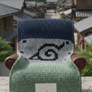 May include: A handmade, knitted blanket in shades of navy blue, gray, and green, draped over a brown leather armchair. The blanket features a black and gray graphic design. The background shows a village scene with mountains.