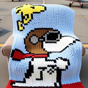 May include: A light blue knitted blanket featuring a pixelated image of a cartoon character wearing a pilot's cap and scarf, with a yellow graphic in the upper left corner. The blanket has a red border and is draped over a chair. The background shows airplanes on a runway.