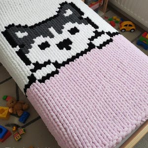 Può includere: Una coperta fatta a mano, a maglia grossa, bianca, grigia, nera e rosa. La parte superiore presenta un disegno a pixel di un muso di cane in bianco, grigio e nero. La metà inferiore è rosa tinta unita. La coperta è su un letto per bambini.