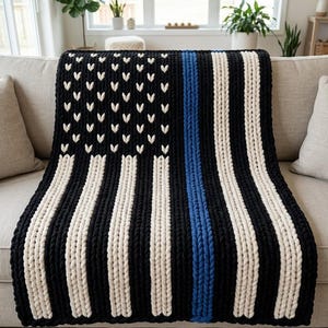 May include: A chunky knit throw blanket featuring a black and white striped pattern with a single vertical blue stripe. The upper left section displays a field of black with white V-shaped stitches arranged in a grid to resemble stars.