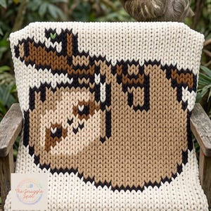 May include: A knitted chair cover featuring a sloth design in shades of brown, black, and cream. The sloth is hanging from a branch, with a real sloth in the background. The chair cover is on a wooden chair.