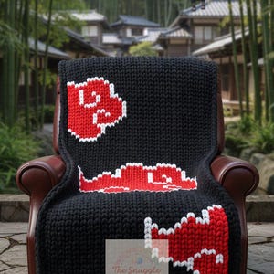 Può includere: Coperta lavorata a maglia nera con disegni di nuvole rosse e bianche. La coperta è drappeggiata su una poltrona in pelle marrone. Il logo The Snuggle Spot è visibile. Lo sfondo presenta un villaggio in stile giapponese.