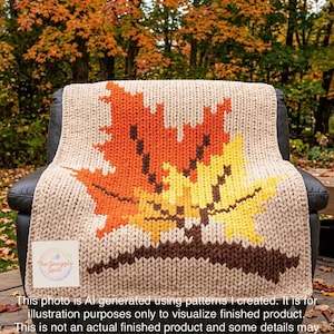 Op de afbeelding: Een grof gebreide deken in beige met een groot, centraal motief van twee esdoornbladeren in oranje en geel met een bruine steel. Een vierkant wit label in de linkerbenedenhoek bevat de tekst The Snuggle Spot.