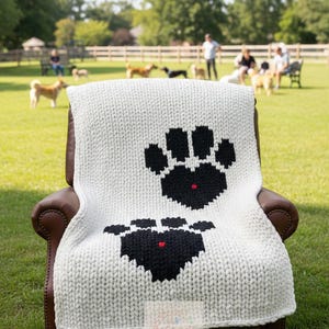 May include: A white knitted blanket with two large black paw prints and red accents, draped over a brown leather armchair. The blanket is on a green lawn with dogs and people in the background. The text on the image says, "This photo is AI generated and is for illustration purposes only to visualize finished product. This is not an actual finished product."