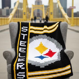 May include: A black, yellow, and white crocheted blanket with the word "STEELERS" vertically displayed. The blanket features the team's logo in the centre, with a yellow, red, and blue diamond shape. The blanket is draped over a grey chair.