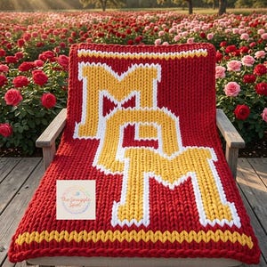 May include: A red knitted blanket with the letters "M" and "M" in yellow and white. The blanket is draped over a wooden chair in a rose garden. The blanket has a yellow and white border. The text "The Snuggle Spot" is in the corner.