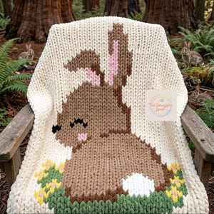 May include: A cream-coloured, chunky knit blanket with a brown bunny design, pink ears, and a white tail. The bunny is set against a green and yellow floral pattern. The text on the blanket reads: "The Snuggle Spot".