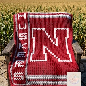 May include: A red and white knitted blanket with a large white "N" and the word "HUSKERS" in white. The blanket is draped over a wooden chair. The background features a field and a red barn.