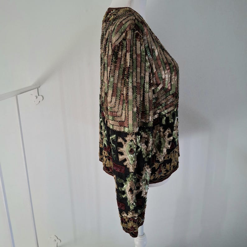 Puede incluir: Top de manga larga con lentejuelas con un patr&oacute;n geom&eacute;trico en tonos marr&oacute;n, verde y dorado. La parte inferior del top presenta un fondo negro con un dise&ntilde;o floral. El top se exhibe en un maniqu&iacute; blanco.