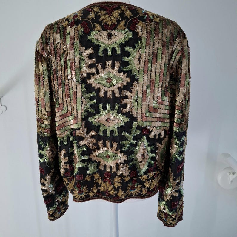 Puede incluir: Una chaqueta negra con intrincados detalles de lentejuelas. La chaqueta presenta un patr&oacute;n geom&eacute;trico en tonos verdes, dorados y marrones. Las lentejuelas crean un efecto texturizado y brillante, agregando un toque de glamour a la prenda.