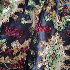 Puede incluir: Primer plano de una tela oscura adornada con lentejuelas y cuentas de colores. El dise&ntilde;o presenta patrones intrincados en tonos de verde, dorado, rojo y crema. Las lentejuelas est&aacute;n cosidas a la tela, creando una apariencia texturizada y ornamentada.