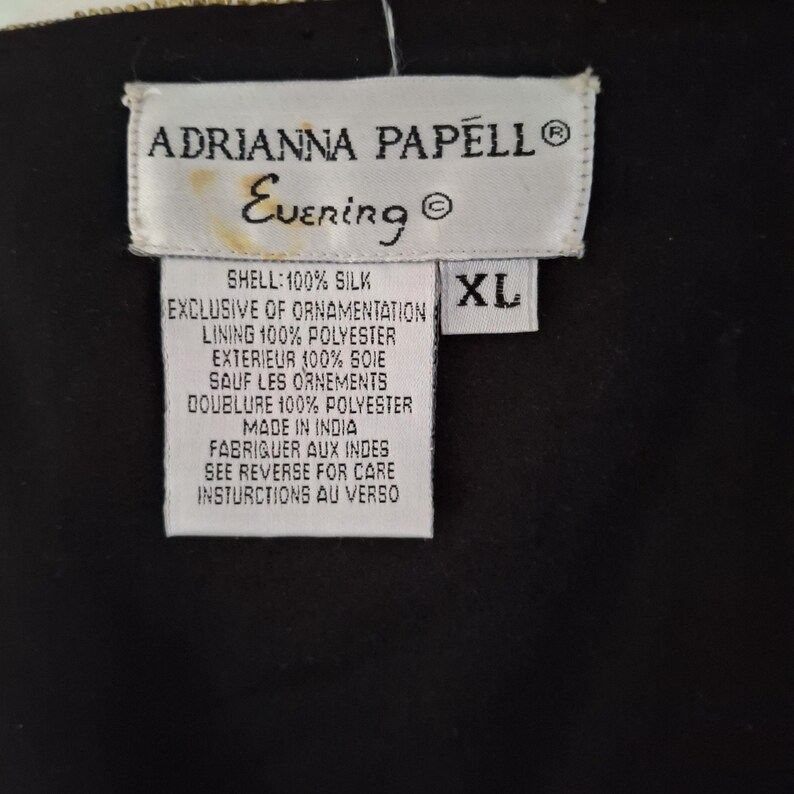 Puede incluir: Primer plano de una etiqueta blanca con el texto "ADRIANNA PAPELL Evening" y detalles de la prenda. La etiqueta tambi&eacute;n indica la talla XL. La prenda est&aacute; hecha en India.