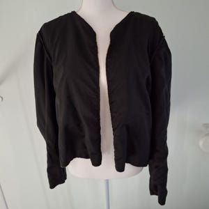 Puede incluir: Una chaqueta negra con mangas largas y frente abierto. La chaqueta se exhibe en un maniqu&iacute; blanco. La tela de la chaqueta parece ser de color liso y uniforme. La chaqueta tiene un dise&ntilde;o de cuello en V.