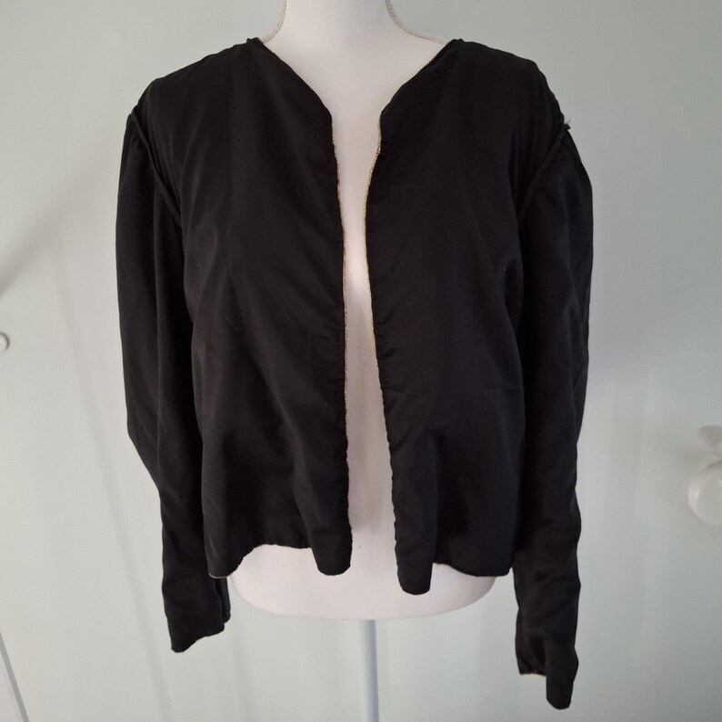 Puede incluir: Una chaqueta negra con mangas largas y frente abierto. La chaqueta se exhibe en un maniqu&iacute; blanco. La tela de la chaqueta parece ser de color liso y uniforme. La chaqueta tiene un dise&ntilde;o de cuello en V.