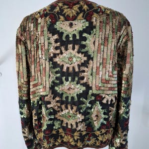 Puede incluir: Una chaqueta negra con intrincados adornos de lentejuelas y cuentas. La chaqueta presenta un patr&oacute;n geom&eacute;trico en tonos dorados, verdes y rojos. Las mangas y la espalda est&aacute;n completamente cubiertas con el dise&ntilde;o decorativo, creando una apariencia texturizada y ornamentada.