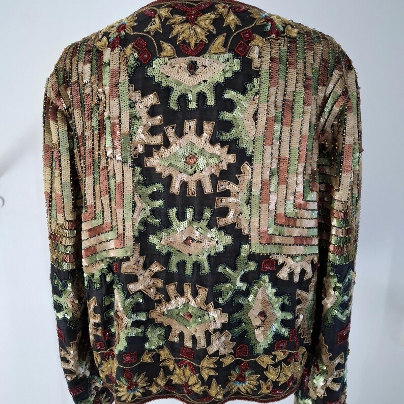 Puede incluir: Una chaqueta negra con intrincados adornos de lentejuelas y cuentas. La chaqueta presenta un patr&oacute;n geom&eacute;trico en tonos dorados, verdes y rojos. Las mangas y la espalda est&aacute;n completamente cubiertas con el dise&ntilde;o decorativo, creando una apariencia texturizada y ornamentada.