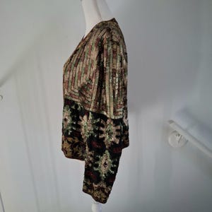 Puede incluir: Chaqueta con lentejuelas y cuello en V. La parte superior presenta rayas verticales en tonos verdes, marrones y dorados. La secci&oacute;n inferior es negra con patrones florales y paisley dorados, verdes y rojos. La chaqueta se exhibe en un maniqu&iacute;.